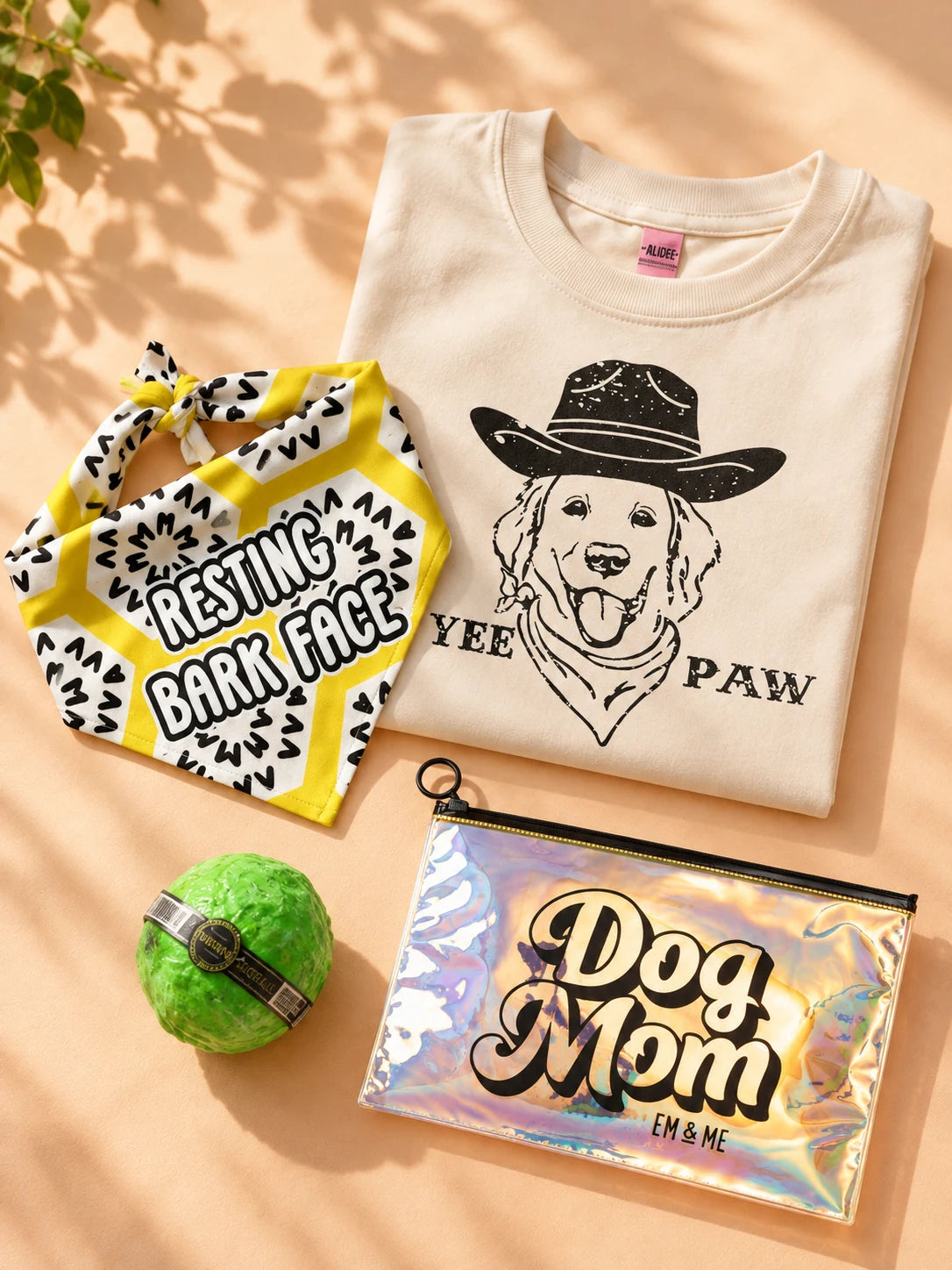 Yee Paw graphic tee, Resting Bark Face bandana, green WunderBall and Dog Mom holographic zipper pouch arranged on a peach tabletop