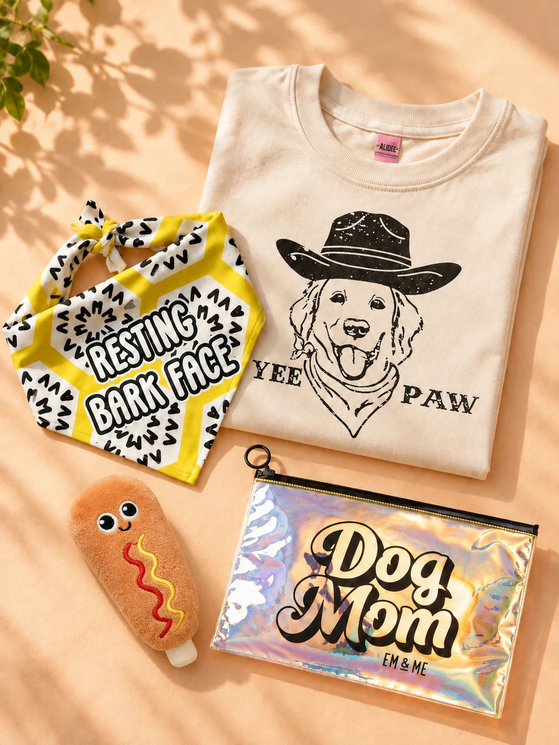 Yee Paw graphic tee, Resting Bark Face bandana, ThoozyPet corndog plush dog toy and Dog Mom holographic zipper pouch arranged on a peach tabletop